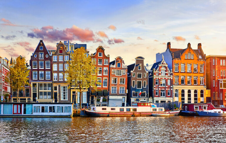 View of Amsterdam from the canal. There are various canal boats docked along the street. Beyond that, there's rows of buildings with different colors (brown, orange, red, yellow). Each building has about 5 floors and they all have the famous pulley at the roof to pull up furniture or large items.