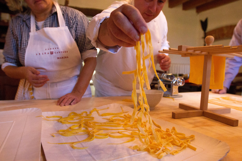 View of two people standing by a kitchen table. Both are dressed like chefs with white apron or outfit. Man on right is holding up home made pasta noodles. Man on left is peering at the noodles.