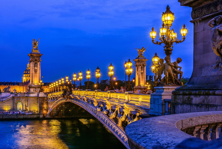 View of Seine River with an old classic bridge spanning it. It's dusk. Bridge has old fashioned lamps spaced across the whole span. There's a monument on the other side with a horse statue on top.