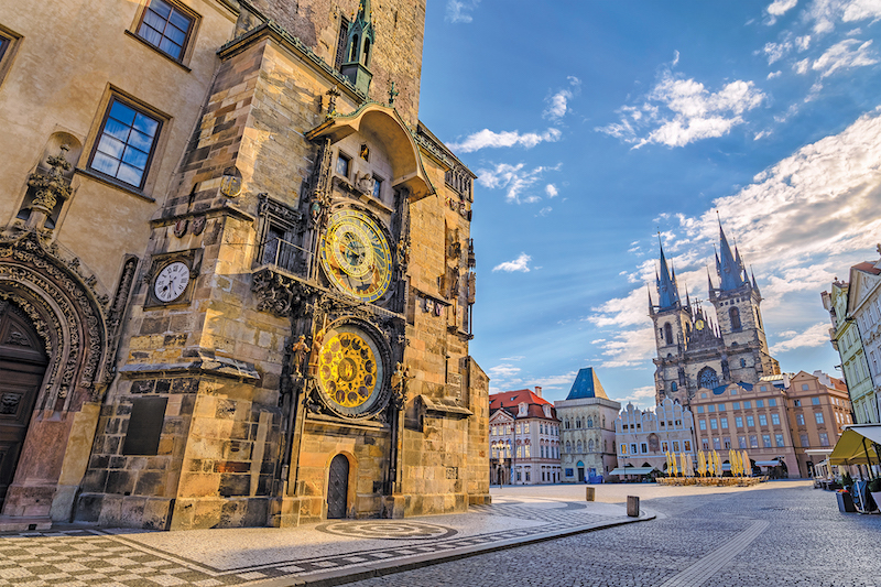 View of a town square in Europe. There's an old building (stone) on left with two giant clocks. One shows the time and the other is a celestial clock. Beyond the church is a view of colorful buildings. Another church dominates the view in the background with two large spires.
