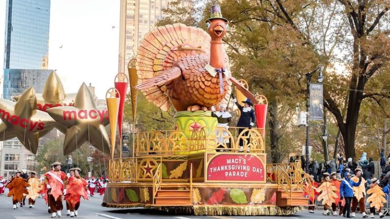 Macy's Thanksgiving Day Parade. A giant inflatable turkey is standing on a float with two people in front. They are dressed like pilgrims.