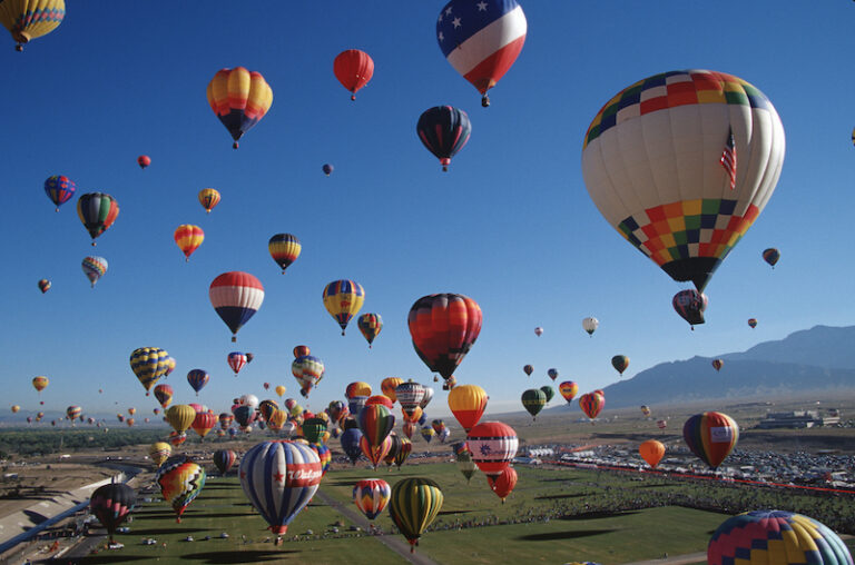New Mexico Balloon Fiesta. View of several giant balloons lifting up from the ground below. There's a mountain range in the background.