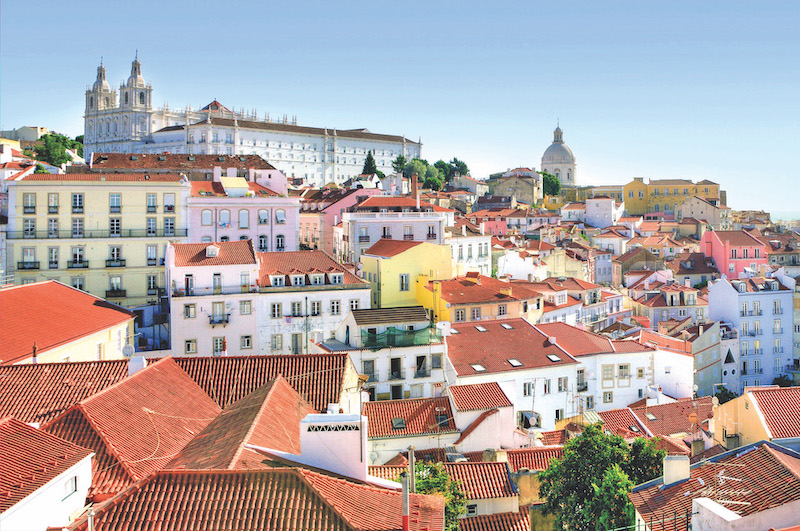View of a large white building at the top of the hill. From that building, there are several buildings going downwards on a slope. Most of the buildings have red tiles. Most buildings are bright white, are about 3 to 6 stories tall.