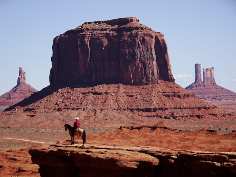 View of a large rock protruding from the ground. Rock is circular and it's quite massive. It's reddish in color. There's a man on a horse at the base staring at it.