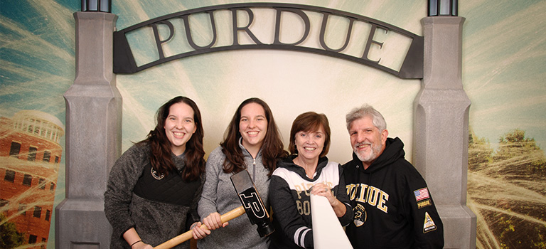 Four cheerful alumni posing for a photo in front of a Purdue archway background at the Boilermaker Station Welcome Center.