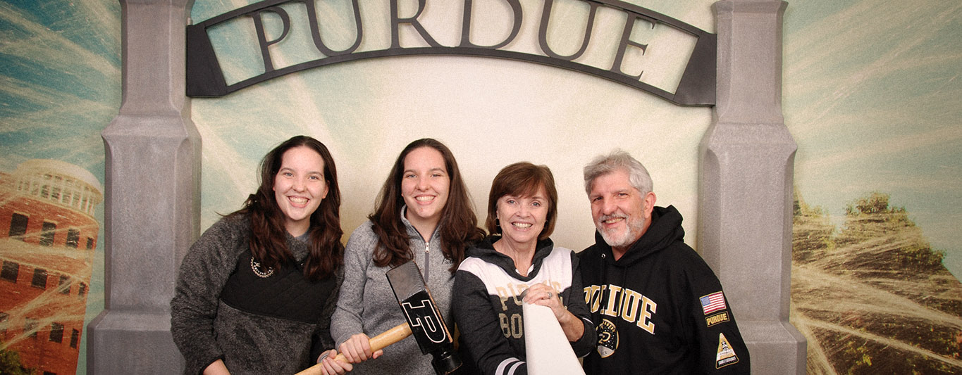 Four people holding a Purdue University hammer in from of Purdue Boilermaker Station Welcome