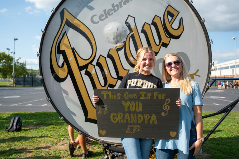 2 women standing in front of Purdue's Big Bass Drum holding a sign that reads "This one is for you grandpa"
