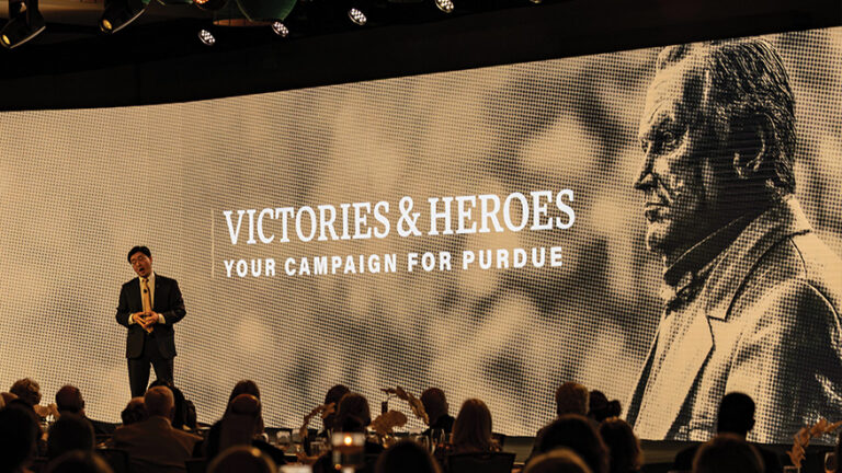Purdue President Mung Chiang standing in front of a giant screen with Victories & Heroes Your Campaign for Purdue logo