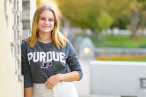 Hannah Majewski, wearing a gray long-sleeved shirt with PURDUE on it, leaning on  a building. 