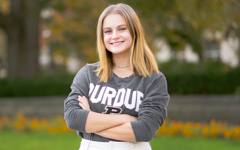 Hannah Majewski, wearing gray long-sleeved shirt with PURDUE on it. She's standing in front of group of trees.