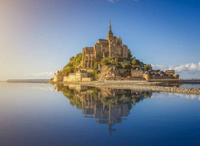 Beautiful panoramic view of famous Le Mont Saint-Michel tidal island with deep blue water and clear reflections in golden evening light at sunset in summer, Normandy, northern France.