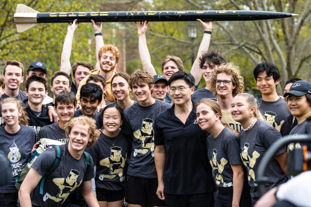 Purdue President Mung Chiang with a group of students smiling. Two students are holding up a model rocket in the background