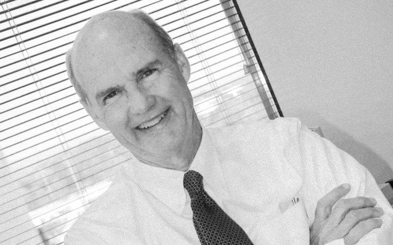 White male with white button-down shirt, black tie, leaning to the photo's left. Background: window with blinds drawn.