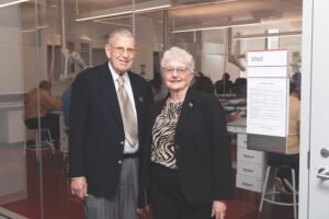 Two people standing in front of glass. Beyond glass is a laboratory with students sitting down. Dave Hale is on the left with a suit and gold tie. Judy Hale is on the right with a jacket and blouse.  