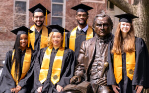 5 Purdue graduates in their caps and gowns standing next to the John Purdue statue