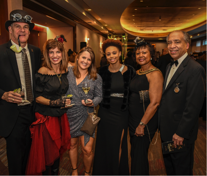 4 women and 2 men dressed in steampunk outfits, tuxedos, and dresses with their arms wrapped around each other smiling