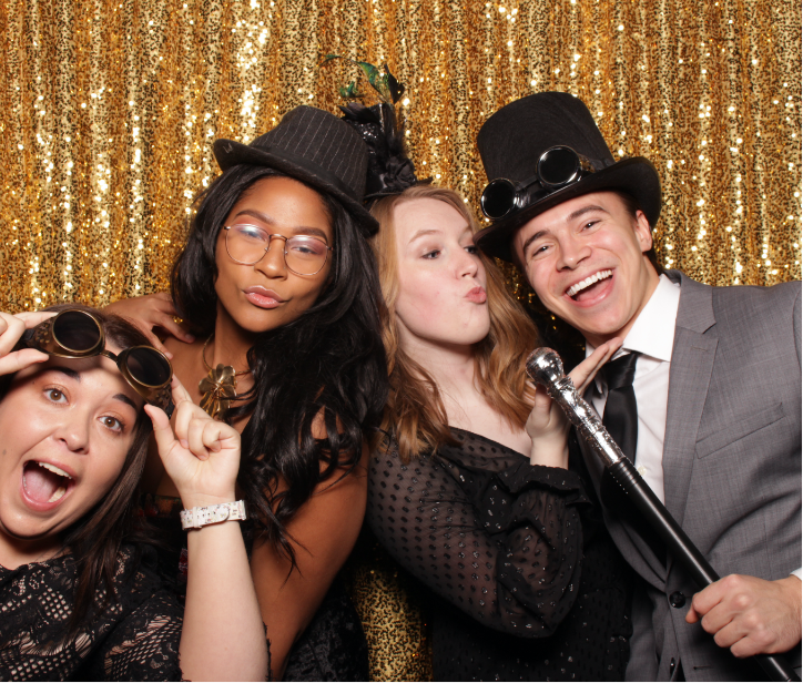 3 Woman and 1 man wearing steampunk outfits posing in front of a gold background smiling