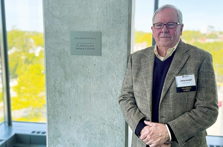 Man with white hair and glasses and tweed jacket standing in front of a plaque. Plaque reads: "Collaboration Space through the generosity of Dr. Jeffrey S. and Barbara E. Kristoff"