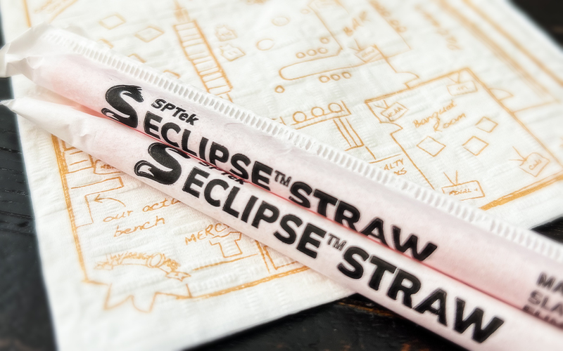 Napkin with blueprint outline in orange of a restaurant. On top of the napkin, two straws with the label: SPTeK Eclipse Straw