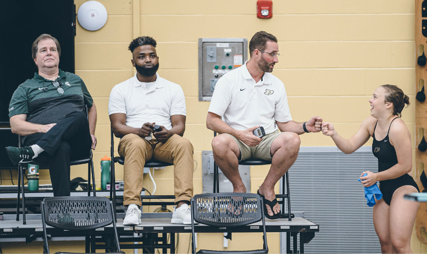 2 men wearing white polos sitting on a stage where one man is fist bumping a Purdue University diver