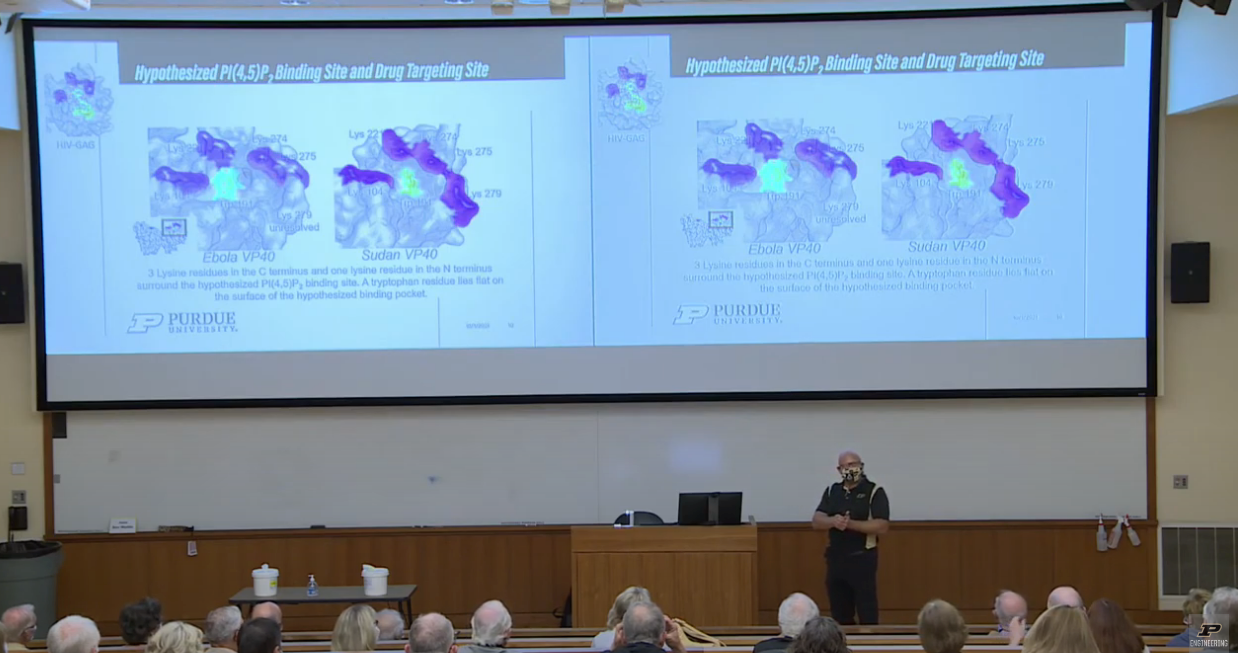 Two projected screens on the wall. Both showing different types of cells and scientific terms. Man is standing in front of the two screens, giving a lecture.